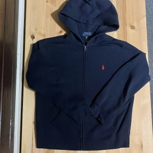 Mörkblå hoodie från Polo Ralph Lauren - Snygg mörkblå hoodie från Polo Ralph Lauren med dragkedja och huva. Klassisk röd broderad logga på bröstet. Tillverkad i mjuk bomull och har långa ärmar. Ny pris är 1100 men priset kan fortfarande diskuteras.