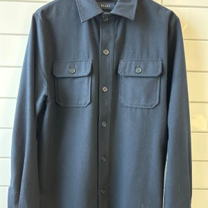 🔥Overshirt från Riley🔥 - Stilren svart overshirt från Riley med klassisk krage och två stora bröstfickor med knappar. Skjortan har långa ärmar och är tillverkad i ett mjukt material som känns skönt mot huden. Perfekt att slänga på sig över en t-shirt för en chill look. I ull så bra till lite kallare väder. Köpt på Brothers för 1600 kr. Har fler bilder och mått är bara att fråga 🔥passar 178-188 📏