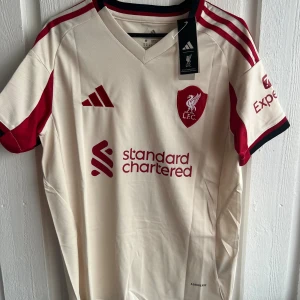 Liverpool Adidas fotbollströja beige/röd - Snygg Liverpool fotbollströja från Adidas i beige med röda detaljer och tryck. Tröjan har korta ärmar, klubbmärke på bröstet, sponsorlogga och Champions League-märke på ärmen. Namn och nummer 'ISAK 9' på ryggen. Tillverkad i lätt och ventilerande material.