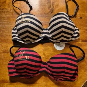 Två nya randiga Snoopy-behåar 75B - Två behåar från Snoopy, en i svart och beige med guldtryck samt en i svart och röd med guldtryck. Båda har horisontella ränder och motiv av Snoopy på kupan. Materialet är mjuk bomull med elastan för skön passform. Kuporna är tunna och passerade Justerbara tunna axelband och klassisk form. Helt oanvända, den ena har orginallappen kvar. Från konkurslager. Finns många fler varianter och storlekar. Rabatt vid flerköp.