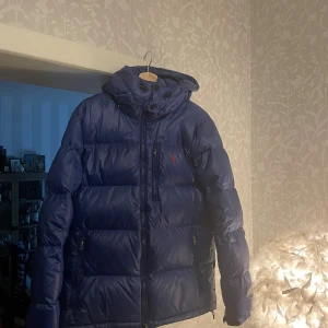Blå dunjacka från Ralph Lauren - Säljer en blå dunjacka från Ralph Lauren med huva och dragkedja. Jackan har en klassisk pufferstil, logga på bröstet och fickor med dragkedja. Perfekt för kalla dagar och snygg till streetwear. Vid smidig affär kan priset såklart diskuteras! Kan skickas fler bilder vid förfrågan 