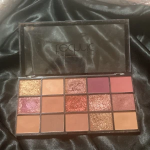 Technic ögonskuggspalett med 15 färger - Säljer en Technic ögonskuggspalett med 15 olika nyanser i både matta och skimrande toner. Färgerna går i guld, koppar, rosa, lila, nude och bruna nyanser. Perfekt för att skapa allt från neutrala till glittriga looks. Paletten har en genomskinlig plastförpackning.