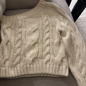 Beige kabelstickad tröja - Säljer en  beige kabelstickad tröja med bred ribbad kant vid hals, ärmslut och nederkant. Tröjan har ett klassiskt flätmönster och är perfekt för kyliga dagar. Den är mjuk och har en relaxed fit med långa ärmar.