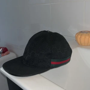 Svart Gucci keps med GG-mönster - Säljer en svart keps från Gucci med diskret GG-monogram över hela kepsen. Klassisk böjd skärm och ikoniskt grönt och rött band på sidan. Snygg och stilren accessoar som lyfter vilken outfit som helst.