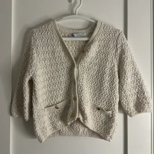 Beige stickad kofta från Zara med guldknappar - Superfin beige stickad kofta från Zara med snygga guldknappar och diskret glittrig tråd. Koftan har trekvartsärm, två små fickor framtill och ett strukturerat mönster som ger en cool vintagekänsla. Perfekt att slänga över en topp för extra stilpoäng.