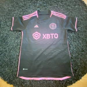 Inter Miami CF svart och rosa matchtröja - Svart fotbollströja från Inter Miami CF med rosa detaljer och Adidas-logga. Tröjan har korta ärmar, rosa ränder på axlarna och klubbmärke på bröstet. Materialet är lätt och andas, Aeroready-teknologi för extra komfort. Snyggt vågmönster på framsidan och sponsortryck.