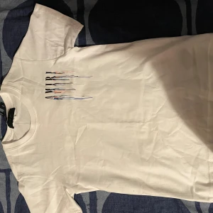 Vit AMIRI t-shirt med färgstänk - Snygg vit t-shirt från AMIRI med coolt färgstänksmönster och stor logga både fram och bak. T-shirten har korta ärmar och är gjord i mjuk bomull. Perfekt för dig som vill sticka ut med en unik och trendig look.