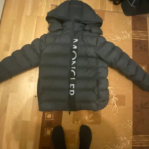 Svart Moncler dunjacka med huva - Säljer en svart dunjacka från Moncler med stor huva och tydlig logga längs dragkedjan. Jackan är quiltad och har en puffig siluett som ger en riktigt fet vinterlook. Perfekt för dig som vill ha både stil och värme under kalla dagar.