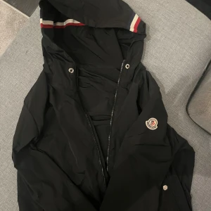 Svart vindjacka från Moncler med huva - Svart vindjacka från Moncler med klassisk logga på ärmen och huva med röd, vit och blå randig insida. Varan är äkta, hör av er vid frågor och funderingar 