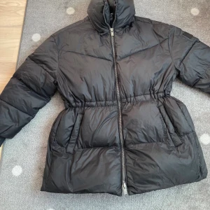 Svart supervarm pufferjacka från Svea, S - Säljer en svart pufferjacka från Svea med hög krage, dragsko i midjan och tvåvägsdragkedja. Jackan är tillverkad i nylon med polyesterfoder och har broderad Svea-logga på huvan. Perfekt för kalla dagar och riktigt snygg passform. Nypriset var  1995:- Supervarm!!