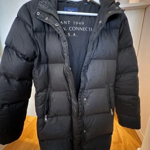 Svart lång pufferjacka från Gant - Svart pufferjacka från Gant med huva och långa ärmar. Jackan har två snedställda fickor med dragkedja och quiltad design. Perfekt för kalla dagar med sin långa modell och stilrena look. Innerfoder med Gant-tryck. 