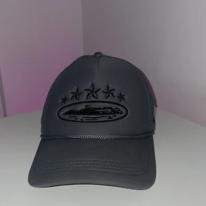 Svart mesh truckerkeps med broderi - Snygg svart truckerkeps med mesh på sidorna och bak, samt justerbar snapback. Framsidan har broderade stjärnor och ett ovalt motiv i svart. Kepsen har en cool streetstil och passar perfekt till en avslappnad look.