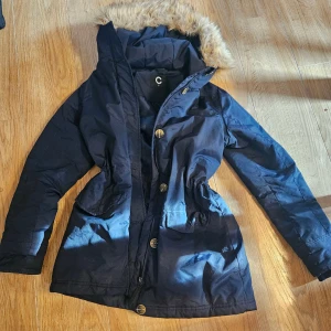 Mörkblå parkajacka med fuskpäls, small  - Snygg mörkblå parkajacka från C med huva och fuskpäls i beige ton. Avtagbar päls i huvan. Jackan har knappar framtill, dragsko i midjan och två stora fickor. Perfekt för kalla dagar med sin långa passform och varma material. Stilsäker look med klassisk design och fluffig krage. Dragkedjan trasig .