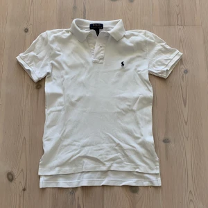 Vit pikétröja från Polo Ralph Lauren - Vit pikétröja från Polo Ralph Lauren med klassisk krage och korta ärmar. Tröjan har den ikoniska broderade loggan på bröstet. Perfekt för en clean och stilren look. Storlek S i barnstorlek