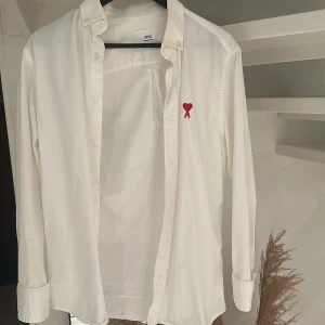 Vit skjorta från Ami med rött hjärta - Vit skjorta från Ami med klassisk krage och långa ärmar. På bröstet finns ett broderat rött hjärta med bokstaven A, som ger en unik och snygg detalj. Skjortan har knappar framtill och är gjord i mjuk bomull som känns skön mot huden.