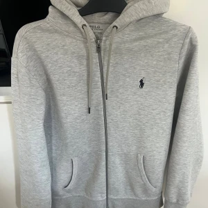 Grå hoodie från Polo Ralph Lauren - Snygg ljusgrå hoodie från Polo Ralph Lauren med dragkedja, huva med snörning och två fickor framtill. Klassisk broderad logga på bröstet. Perfekt för chill dagar och enkel att matcha med jeans eller joggers.