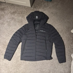 Svart pufferjacka från Sail Racing - Säljer en svart pufferjacka från Sail Racing med huva och dragkedja framtill. Jackan har två sidofickor med dragkedja och diskret logga på bröstet. Den är vadderad och har en stilren, sportig look som passar perfekt när det är kallt ute.