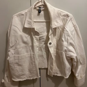 Vit croppad jeansjacka från H&M - Snygg vit croppad jeansjacka från H&M med råa kanter och stora fickor framtill. Jackan har klassisk krage, knäppning med metallknappar och långa ärmar. Perfekt för dig som gillar en avslappnad och trendig stil.