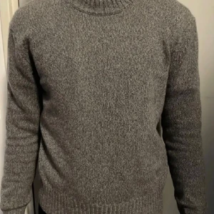 Grå stickad tröja från Ami Paris - Säljer en grå stickad tröja från Ami Paris med diskret broderad logga på bröstet. Tröjan har rund halsringning, ribbade muddar och är långärmad. Perfekt för dig som gillar stilren och klassisk design.