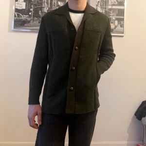 Mörkgrön overshirt med knappar - Stilren mörkgrön overshirt med klassisk krage och stora knappar framtill. Jackan har raka linjer, diskreta fickor på sidorna och är tillverkad i ett mjukt ullmaterial. Perfekt för lager-på-lager och ger en clean look.