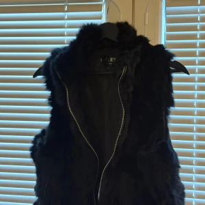 Svart pälsväst från Hollies S - Svart pälsväst från Hollies i storlek S. Västen har fluffig pälslook och en snygg dragkedja framtill. Perfekt för att addera lite edge till din outfit. Fodrad insida och klassisk krage. Enkelt att matcha med jeans eller skinnbyxor.