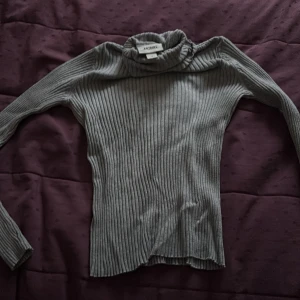 Grå ribbad polotröja från Monki - Säljer en grå ribbad polotröja från Monki i storlek M. Tröjan har lång ärm och en hög, lite rynkad polokrage. Perfekt att styla med jeans eller kjol för en enkel och snygg look.