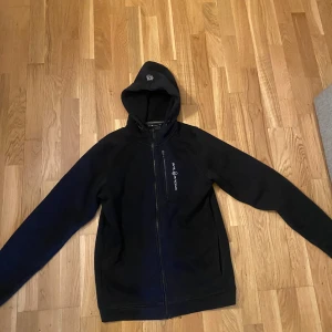 Svart zip hoodie från Sail Racing - Svart zip hoodie från Sail Racing med hel dragkedja och huva. Tröjan har en bröstficka med dragkedja och Sail Racing-logga på bröstet. Mjuk fleece på insidan gör den extra varm och skön även under kalla dagar. Nypris 900kr