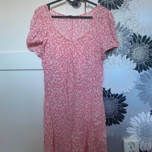 Rosa blommig kortklänning från Cotton On - Supersöt rosa kortklänning från Cotton On med vita små blommor över hela tyget. Klänningen har korta puffärmar, v-ringad hals och knytband i ryggen för extra passform. Perfekt för sommarens alla tillfällen och ger en fräsch och ungdomlig vibe.