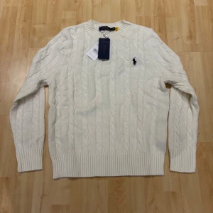 Vit kabelstickad tröja Polo Ralph Lauren - Snygg vit kabelstickad tröja från Polo Ralph Lauren med klassisk rund hals och broderad logga på bröstet. Tröjan har långa ärmar och ribbade muddar vid hals, ärmslut och nederkant. Perfekt för dig som gillar stilrena och tidlösa plagg.