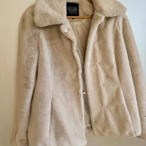Beige pälsjacka från Soul River - Fluffig och mjuk beige (fusk)pälsjacka från Soul River med bred krage och diskret knäppning framtill. Jackan har en clean look och är helfodrad på insidan. Perfekt för dig som vill ha en cozy vibe och gillar neutrala färger.