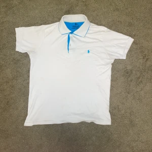 Vit polo från Ralph Lauren, M/S - Vit pikétröja från Polo Ralph Lauren i storlek M men passar S. Klassisk design med blå detaljer på krage och knappslå samt blå broderad logga på bröstet. Tröjan har korta ärmar och är gjord i mjuk bomull som är skön mot huden. Perfekt för en clean och sportig look.