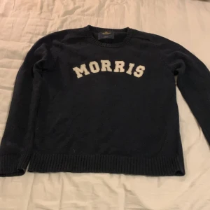 Mörkblå stickad tröja Morris - Snygg mörkblå stickad tröja från Morris med vit text framtill. Klassisk rund hals och ribbade muddar vid ärmslut och nederkant. Perfekt för dig som gillar stilrena college-vibbar och vill ha något bekvämt och tidlöst.