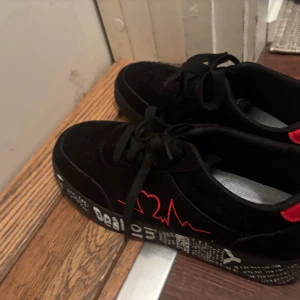 Svarta sneakers med röd detalj - Unika svarta sneakers med coola röda detaljer och grafiskt mönster på sulan. Skorna har snörning, rund tå och är tillverkade i mocka med syntetdetaljer. Röd hjärtpulsbroderi på sidan och texttryck på sulan ger en edgy vibe. Perfekt för dig som gillar streetstyle.