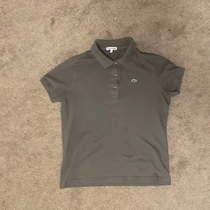 Mörkgrön pikétröja från Lacoste - Säljer en klassisk mörkgrön pikétröja från Lacoste med den ikoniska gröna krokodilen broderad på bröstet. Tröjan har korta ärmar, krage och knappar framtill. Perfekt för en clean och sportig look.