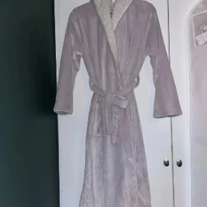 Lila morgonrock  - Mysig ljuslila morgonrock med huva och knytskärp i midjan. Tillverkad i mjukt material med fluffigt foder på insidan av huvan. 