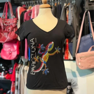 Svart t-shirt med ödla - Svart t-shirt från JHK med färgglad mosaiködla och texten 'Barcelona Gaudí' på framsidan. T-shirten har v-ringning och korta ärmar. 