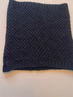 Svart stickad halsduk/tube - Säljer en svart stickad halsduk/tube med grov struktur och ribbad kant upptill och nedtill. Perfekt när det är kallt. Materialet är mjukt och värmande, passar dig som gillar klassisk stil.