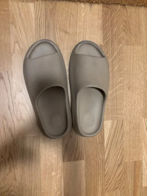 Beige Yeezy Slide sandaler - Säljer ett par Yeezy Slide sandaler i beige färg med chunky, räfflad sula och öppen tå. De är tillverkade i syntetmaterial och har en clean, minimalistisk design som är riktigt populär just nu. Perfekt för chill dagar och enkel att slänga på sig.