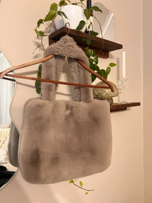 Beige/grå fluffig handväska i päls - Supermjuk och fluffig beige handväska med breda handtag, perfekt för att ge din outfit en mysig vibe. Väskan är rektangulär och har ett stilrent, minimalistiskt utseende. Materialet är pälsimitation som känns lyxigt och trendigt.