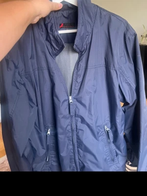 Mörkblå vindjacka från Dressmann - Snygg mörkblå vindjacka från Dressmann i storlek L. Jackan har dragkedja framtill, två praktiska fickor med dragkedja och en hög krage. Materialet är lätt och syntetiskt, perfekt för blåsiga dagar. Enkel och stilren design som funkar till många outfits.