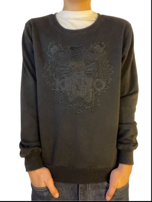 Svart sweatshirt från Kenzo med tiger - Svart sweatshirt från Kenzo med ikonisk broderad tiger och text på bröstet. Tröjan har rund halsringning, ribbade muddar och är långärmad. Tillverkad i mjuk bomull för skön känsla och stilren look.