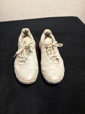 Adidas vita tennis sneakers med röd detalj - Säljer ett par vita Adidas sneakers med röda och silvriga detaljer på sidorna och sulan. Skorna har klassisk snörning, mesh-överdel och tre vita ränder på sidan. Yttersulan har ett mönstrat grepp och är förstärkt med röd och silverfärgad plast. Perfekta för tennis och sport.