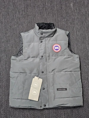 Helt ny Canada Goose väst - Säljer en helt ny grå  från Canada Goose med klassisk hög krage och tryckknappar framtill. Västen har två stora fickor och ett broderat märke på bröstet. Materialet är slitstark polyester och dunfyllning, perfekt för kyliga dagar.