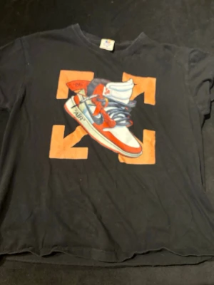 Svart t-shirt med sneaker-tryck - Svart t-shirt från Four Square i 100% bomull. Framsidan har ett stort tryck med en färgglad sneaker och orange grafiska detaljer. Klassisk passform med rund hals och korta ärmar. Perfekt för dig som gillar streetwear och sneakers.