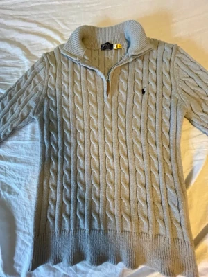 Grå stickad half zip tröja Polo Ralph Lauren - Säljer en grå stickad tröja från Polo Ralph Lauren med half zip och kabelstickat mönster. Tröjan har hög krage, lång ärm och klassisk Polo-logga broderad på bröstet. Perfekt för dig som gillar stilrena och mysiga plagg.