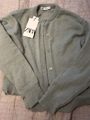 Grön stickad kofta från Zara - Snygg grön stickad kofta från Zara i storlek M. Koftan har rund hals, långa ärmar och knäppning med matchande knappar framtill. Mjuk och skön kvalitet som passar perfekt till lager-på-lager. Enkelt och stilrent plagg för din garderob.