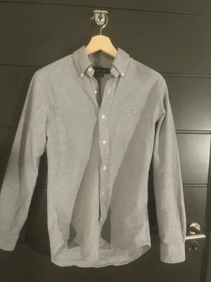 Grå slim fit skjorta från Ralph Lauren - Säljer en grå stretch oxford-skjorta från Ralph Lauren i slim fit. Skjortan har klassisk button-down krage, vita knappar och det ikoniska broderade Polo-logot på bröstet. Långärmad och tillverkad i bomull med snygg, stilren look. Skulle säga att den passar xs Samt s