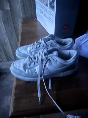 Nike Dunk Low vita sneakers - Säljer ett par Nike Dunk Low sneakers i helvitt. Skorna har klassisk snörning, rund tå och platt sula. Tillverkade i skinn och syntet med perforerade detaljer på tån för extra ventilation. Perfekt för dig som gillar stilrena och ikoniska sneakers.