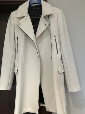 Vit kappa från Zara Woman, storlek M - Säljer en stilren vit kappa från Zara Woman i storlek M. Jackan har snygga silvriga dragkedjor, bred krage och fickor med lock. Materialet känns mjukt och har en ullig look. Perfekt för dig som gillar minimalistisk och clean stil.