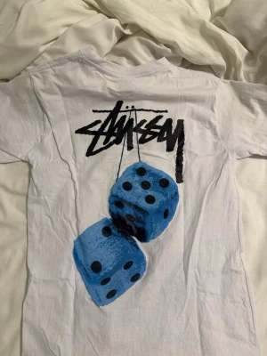 Vit Stüssy t-shirt med blå tärningar - Vit t-shirt från Stüssy med klassisk logga tryckt på bröstet och ett stort grafiskt tryck med två blå tärningar på ryggen. T-shirten har rund hals och korta ärmar, tillverkad i mjuk bomull. Perfekt för dig som gillar streetwear och vill sticka ut med cool design.